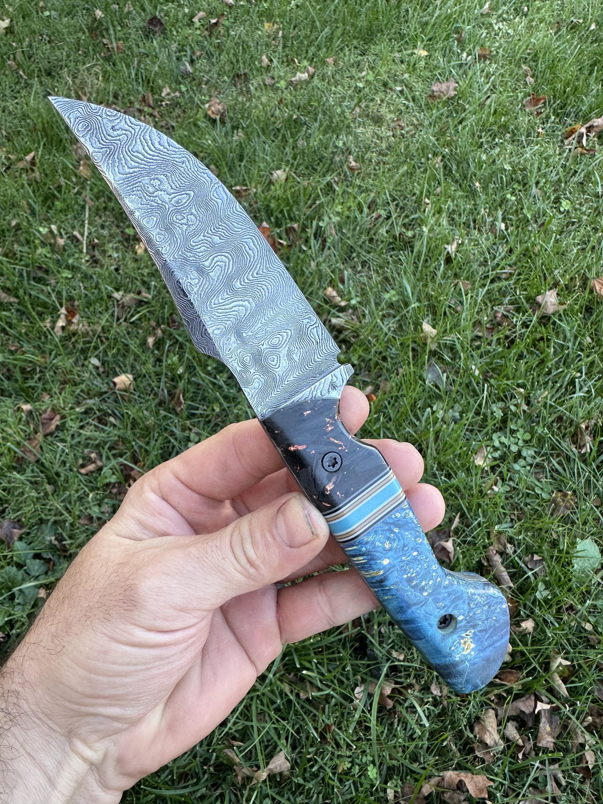 This wicked piece is in a class of its own! With a 130 layer raindrop damscus steel forged in house but with a shimmering super deep etch with a texture you can feel! Handle material is awesome set of combo scales featuring shredded carbon fiber and copper flake with a blue dyed box elder burl wood with a blue glow spacer! (Comes with a kydex sheath) 4.5” blade length 9.5” overall length